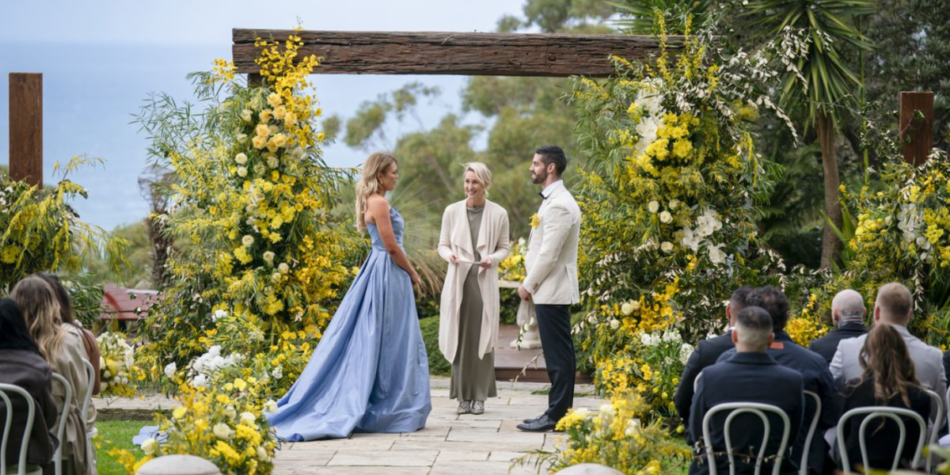 Bride Jacqui Burfoot wears a blue gown, and Ryan Donnelly wears a white tuxedo at their MAFS Australia 2025 wedding, set outdoors with ocean views and a floral arch