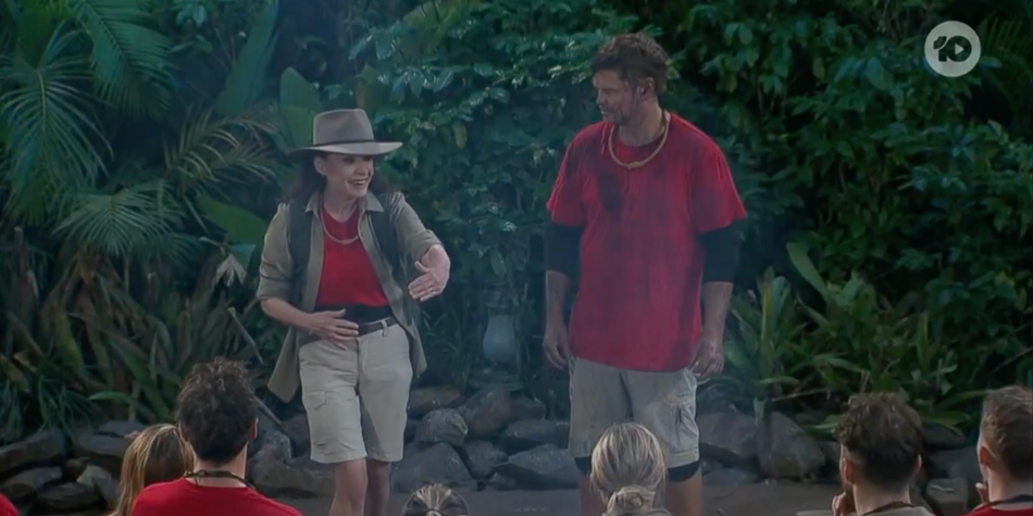 Sigrid Thornton, wearing a wide-brimmed hat and safari-style outfit, and Harrison Reid in a red shirt and shorts, addressing fellow contestants in the jungle camp on I’m a Celebrity 2025