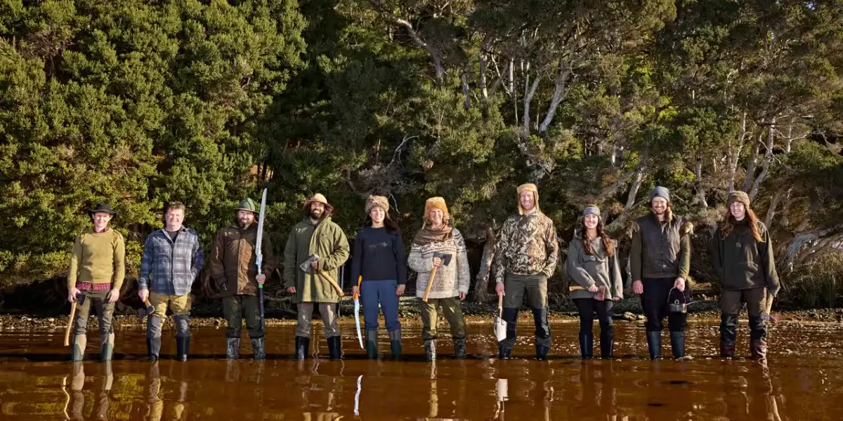 A group photo of the Alone Australia Season 3 cast standing in shallow water, dressed in outdoor survival gear, with a forested background