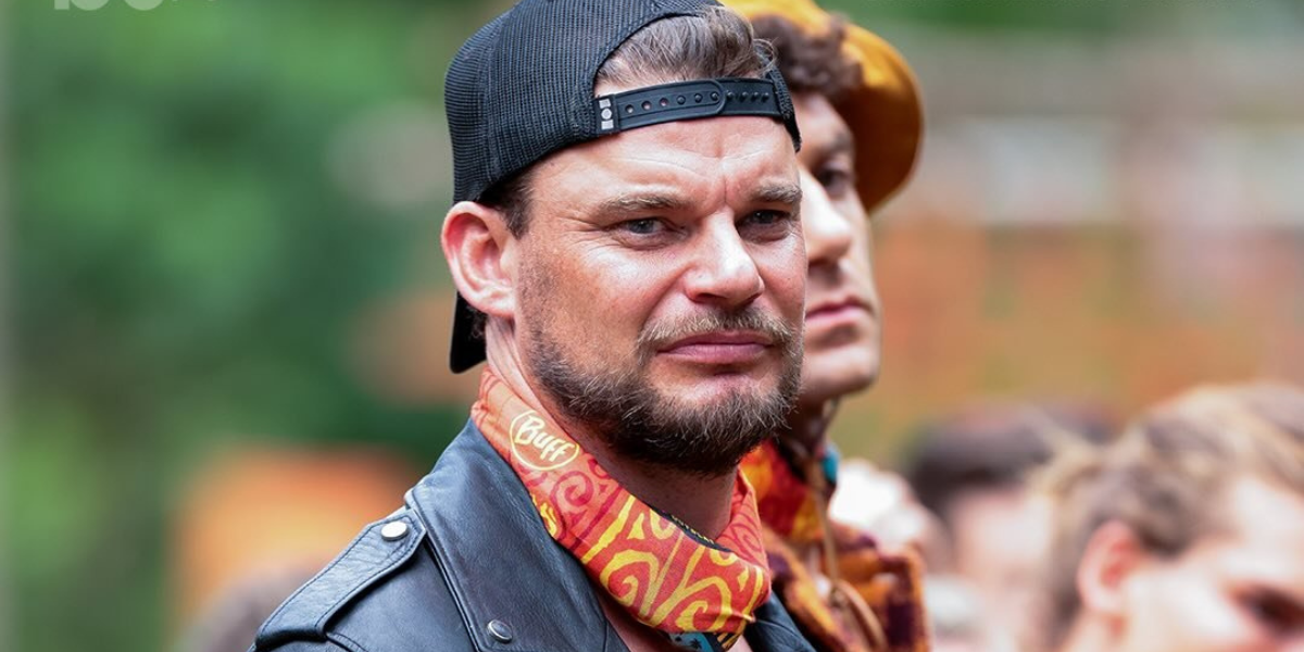 Rich Hughes, wearing a backward black cap, a leather jacket, and a colorful Buff neck gaiter, stands with a serious expression during a challenge on Australian Survivor 2025