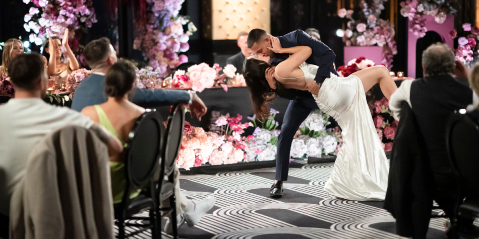 Beth Kelly and Teejay Halkias from Married at First Sight 2025 share a dramatic dip kiss at their wedding reception, surrounded by cheering guests and a lavish floral backdrop