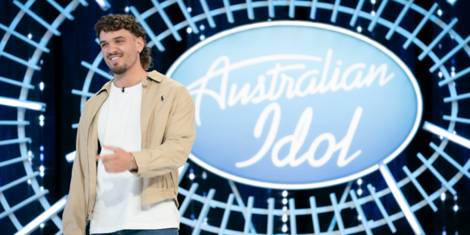 Clint Posselt smiles while auditioning on the Australian Idol 2025 stage, wearing a beige jacket over a white t-shirt with blue jeans