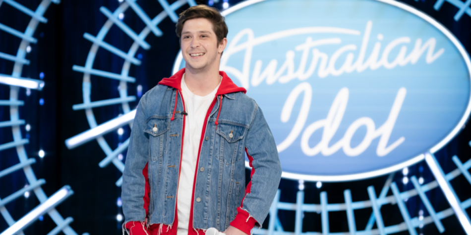 Dinley Jones smiles while auditioning on the Australian Idol 2025 stage, wearing a denim jacket with red hoodie accents over a white t-shirt and holding a microphone