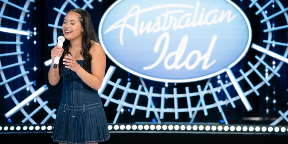 Izellah Connelly sings passionately while auditioning on the Australian Idol 2025 stage, wearing a denim dress and holding a microphone