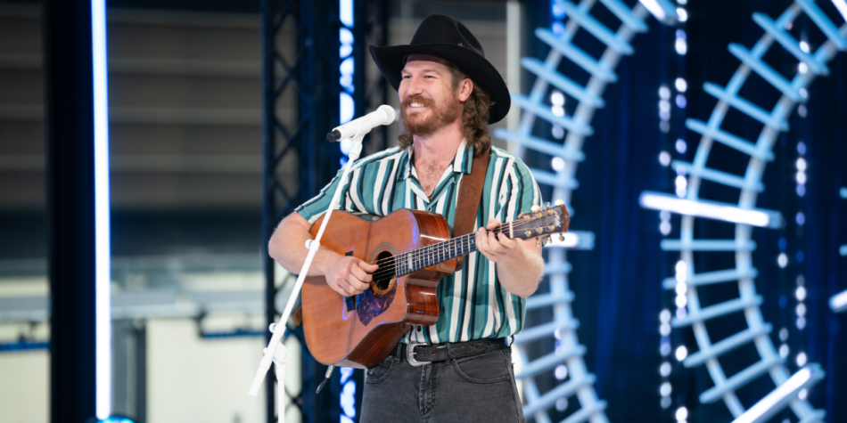 Jake Whittaker performs while auditioning on the Australian Idol 2025 stage, wearing a black cowboy hat, a green and white striped shirt, and playing an acoustic guitar