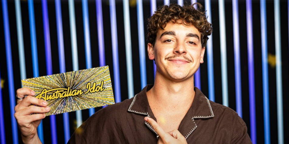 Jaymon Bob smiles while holding his Golden Ticket at the Australian Idol 2025 auditions, wearing a brown short-sleeve button-up shirt against a backdrop of vertical blue and yellow lights