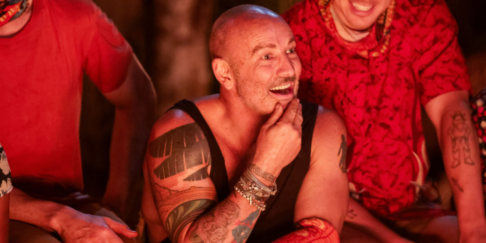 Kent Miller-Randle in a black tank top and bracelets smiles while sitting among fellow contestants around a fire-lit setting on Australian Survivor 2025.