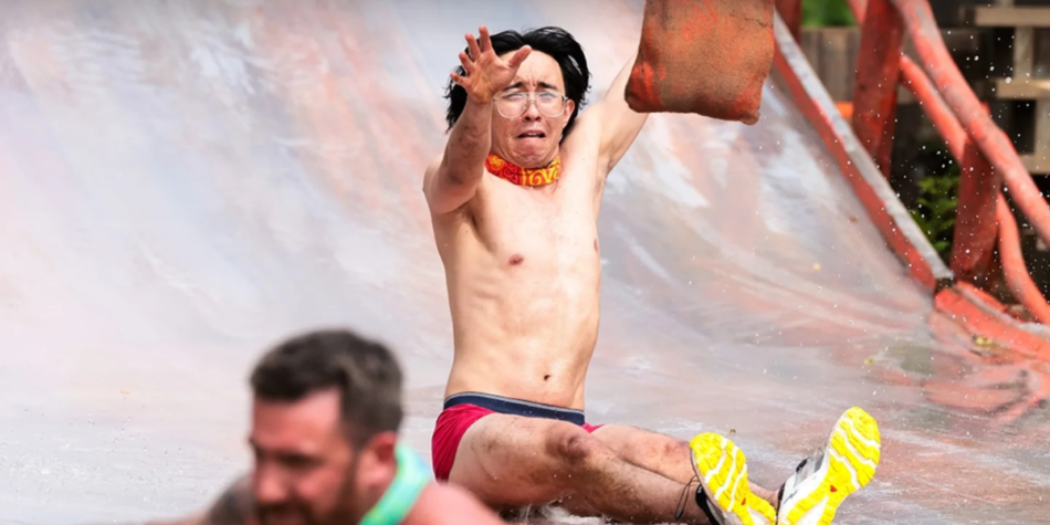 Myles Kuah, wearing glasses, a red buff, and red shorts, slides down a wet ramp with an intense expression, reaching for a sandbag during a challenge on Australian Survivor 2025.