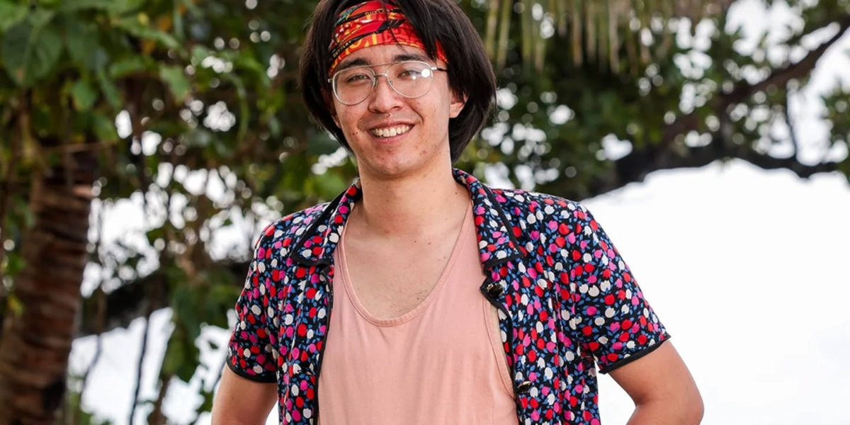 Myles Kuah, wearing glasses, a red buff, and a colorful patterned shirt, smiles while standing outdoors in a tropical setting on Australian Survivor 2025.