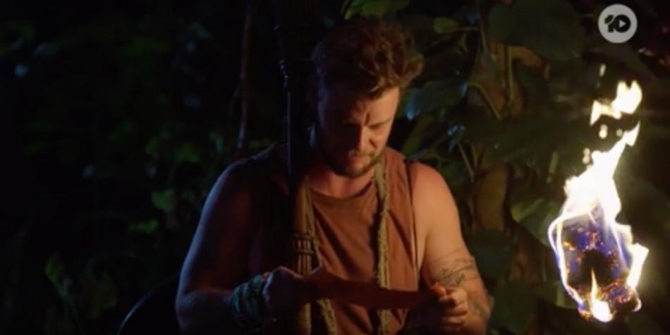 Rich Hughes from Australian Survivor reads a parchment at night while a torch burns brightly beside him.