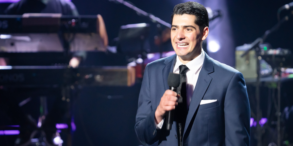 William on stage during Australian Idol 2025, wearing a navy blue suit and holding a microphone while speaking to the audience